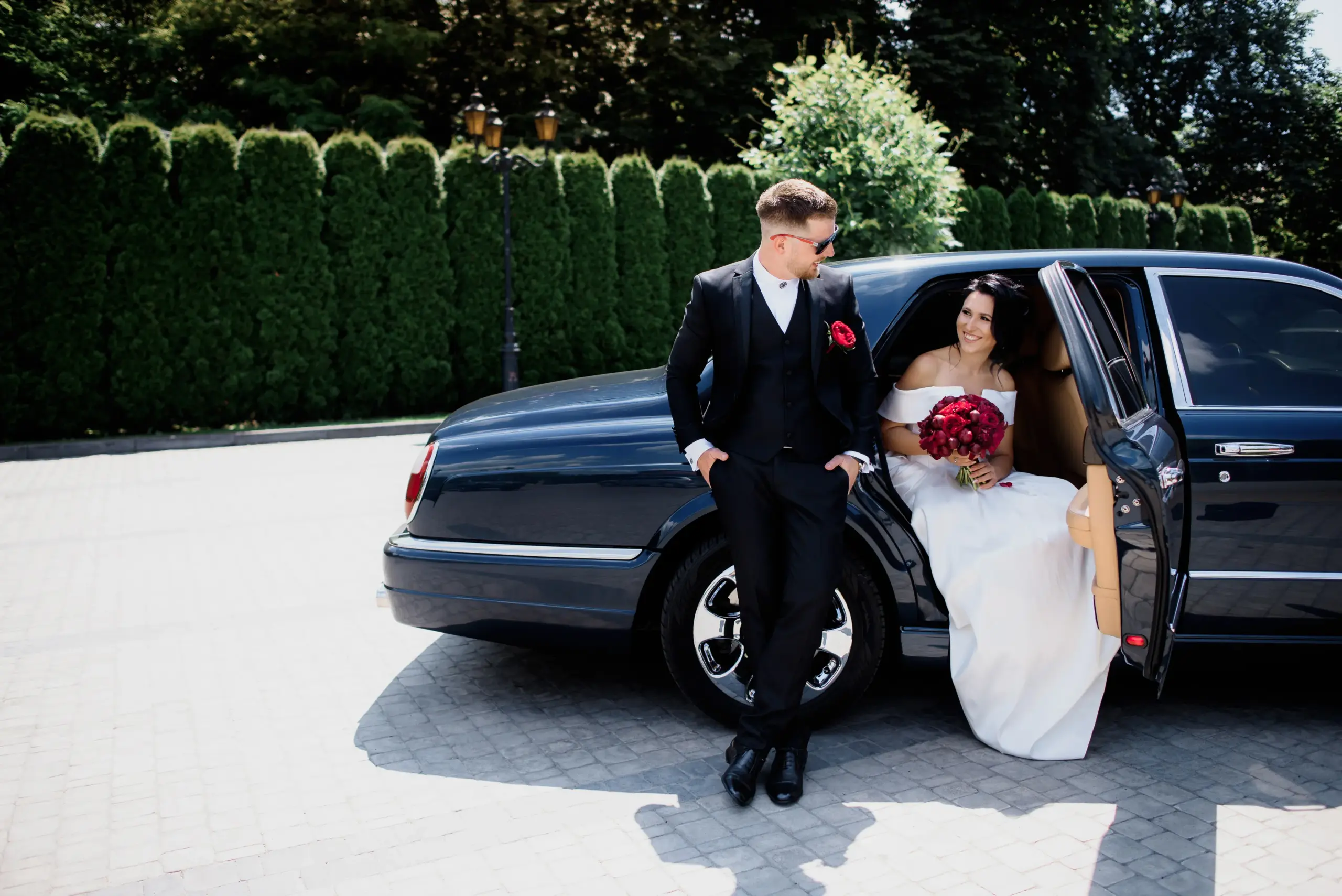 beautiful-wedding-couple-is-smiling-black-car-sunny-day-dressed-elegant-wedding-outfits-with-red-bouquet