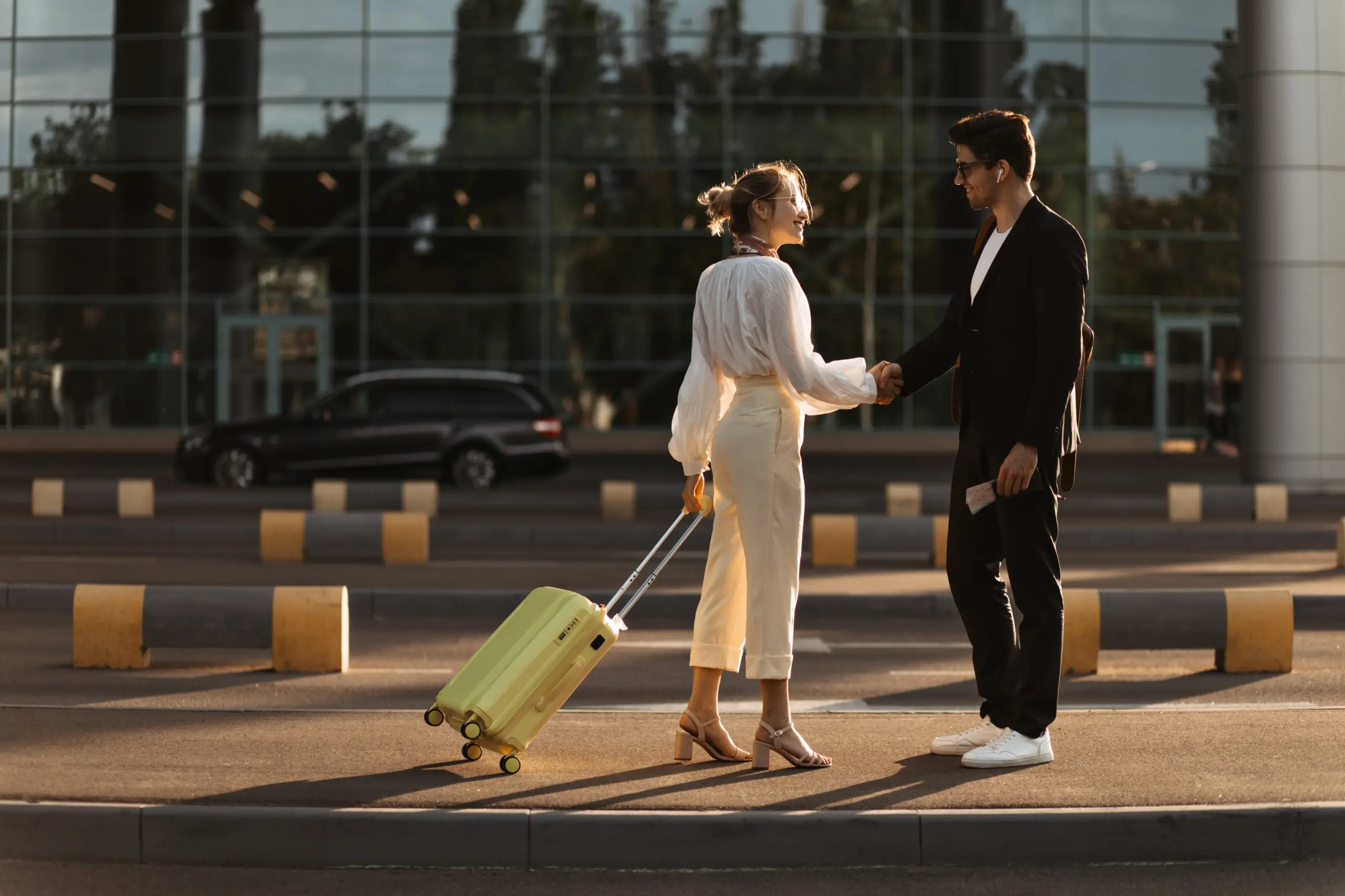 happy-blonde-woman-white-blouse-beige-pants-holds-yellow-suitcase-greets-with-man-black-suit-near-airport